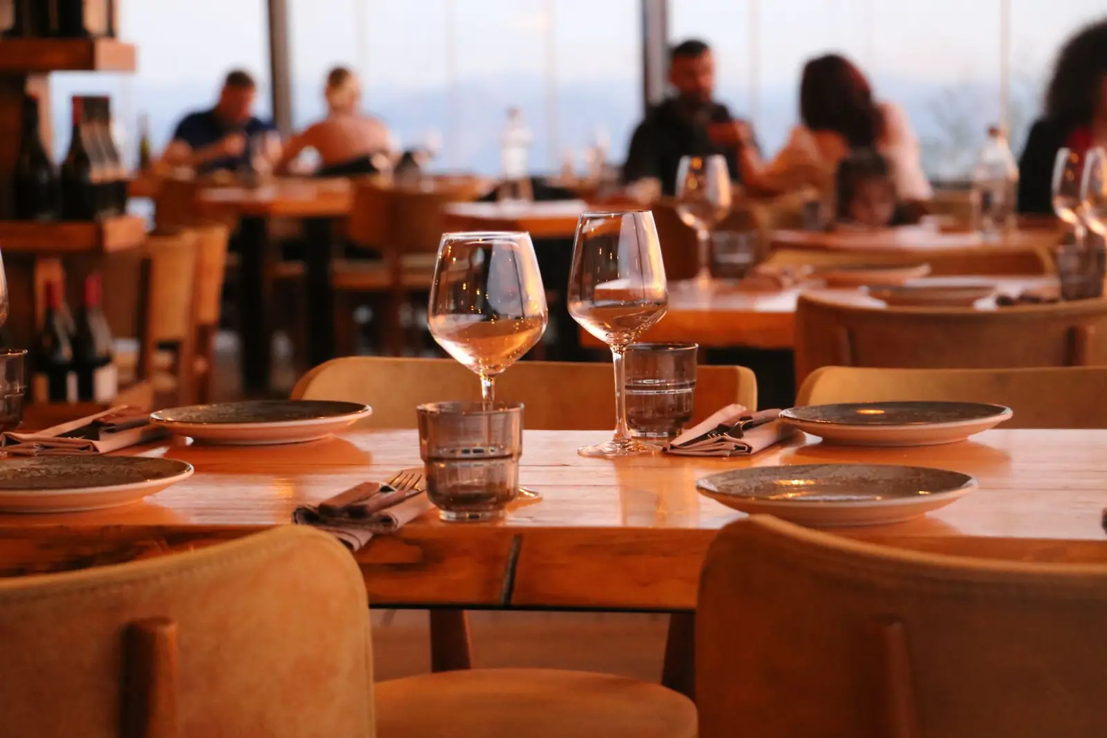 Set wooden tables with wine glasses in a warm restaurant, guests dining by the windows behind